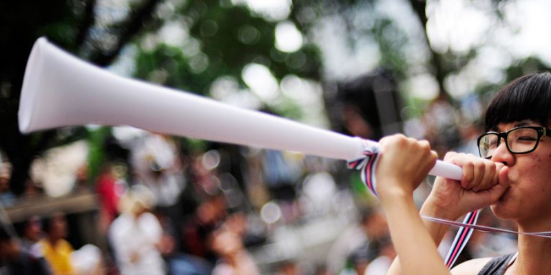 Asian woman sounding a white vuvuzela on a blurred crowd background